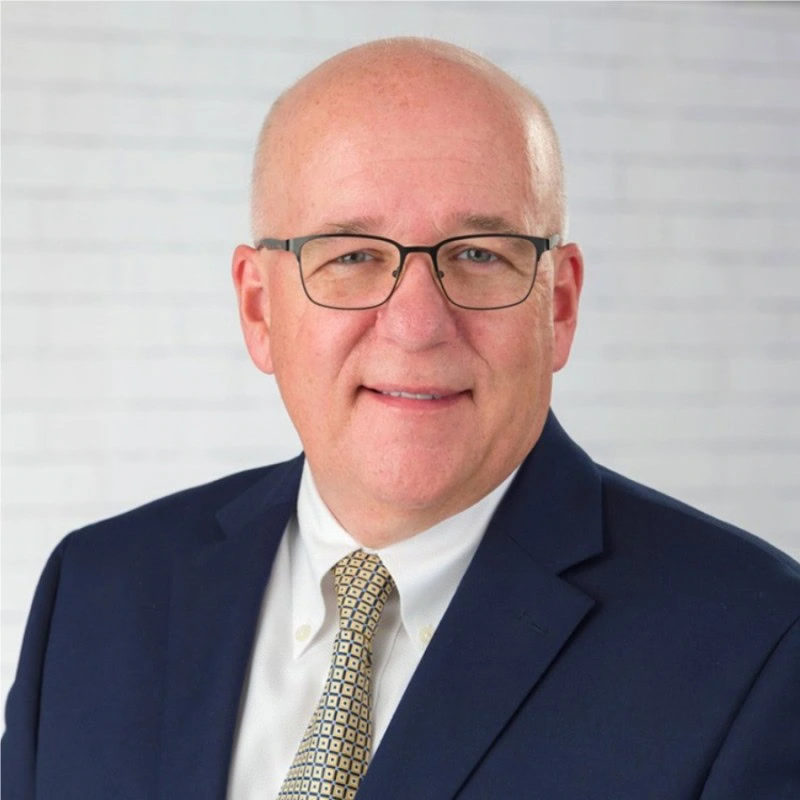 A middle-aged man with glasses, wearing a navy blue suit, white shirt, and patterned tie, poses in front of a white brick background, appearing ready to deliver a Message From the President.