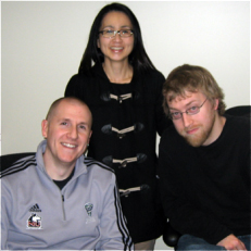 Three people pose indoors, two sitting and one standing behind them, all smiling or looking at the camera against a plain backgroundโperfect for showcasing a friendly helpdesk team.