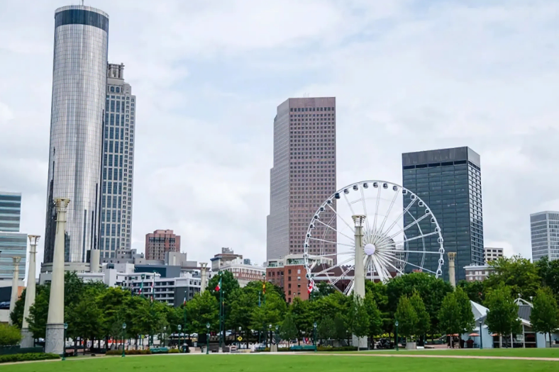 Cityscape view featuring tall buildings, a Ferris wheel, and green trees in the foreground under a cloudy sky.