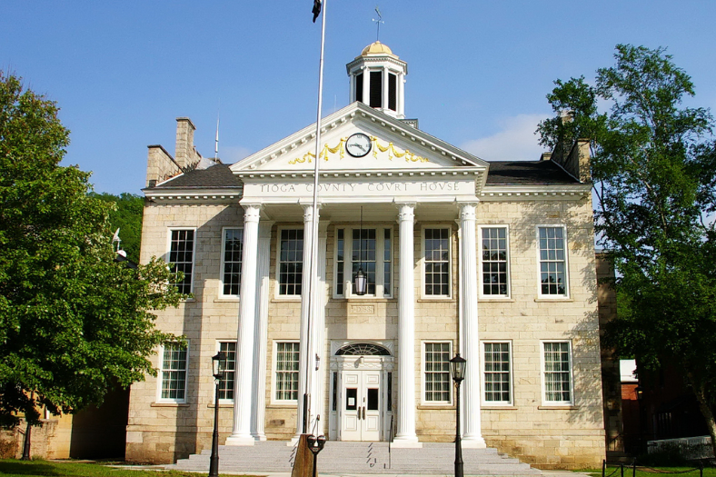 Historic stone courthouse with large white columns, a clock, and cupola on the roof, set against a blue sky and surrounded by trees.